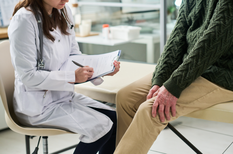 Man in a medical room holding their knee, with a female practitioner with a clipboard